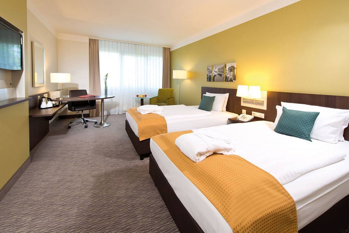 Ein Hotelzimmer mit zwei Betten, einem Schreibtisch, einem Stuhl, einer Lampe und einem Fernseher.