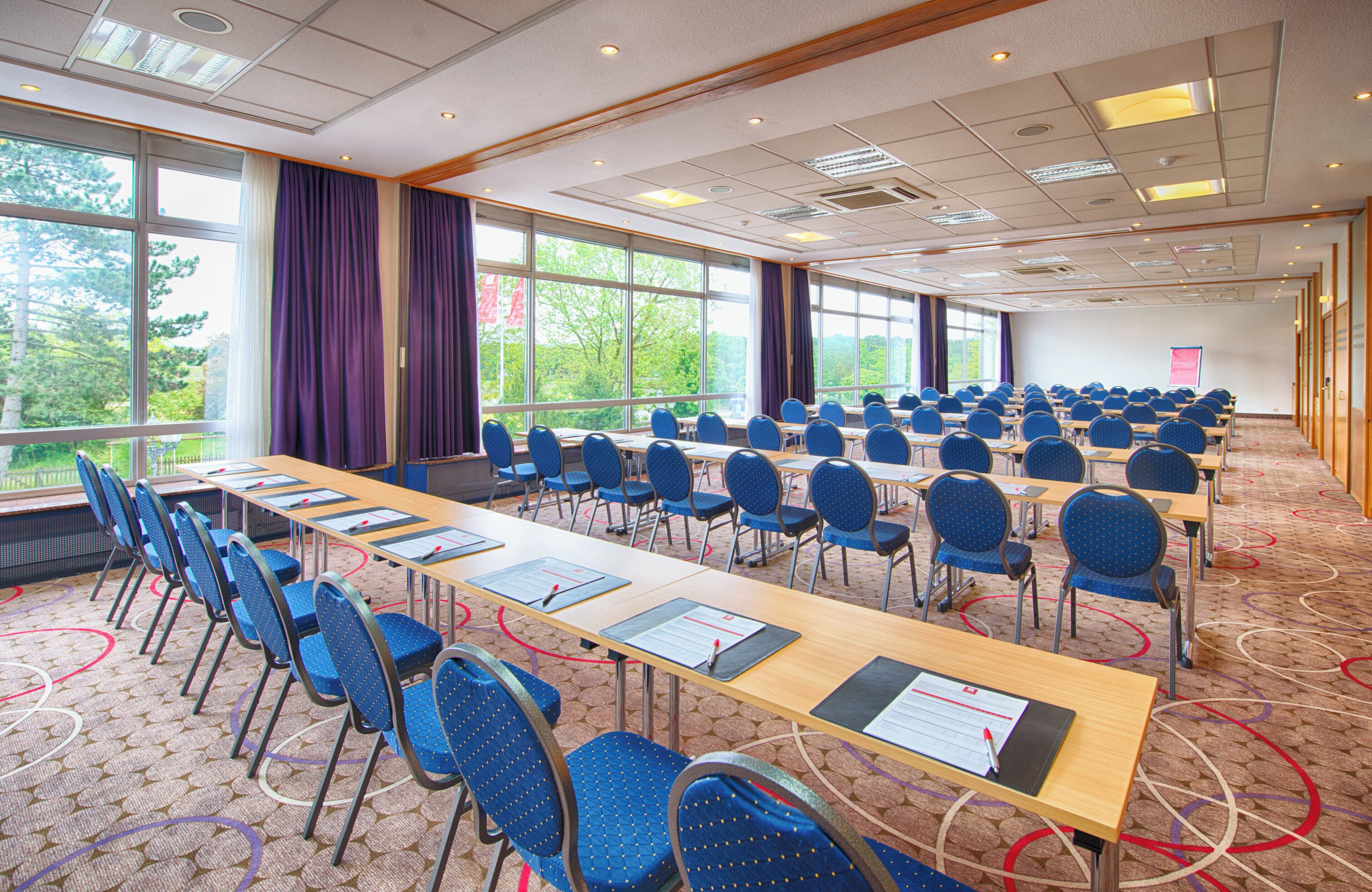 Una sala de conferencias con filas de mesas y sillas, ventanas amplias y suelo alfombrado.
