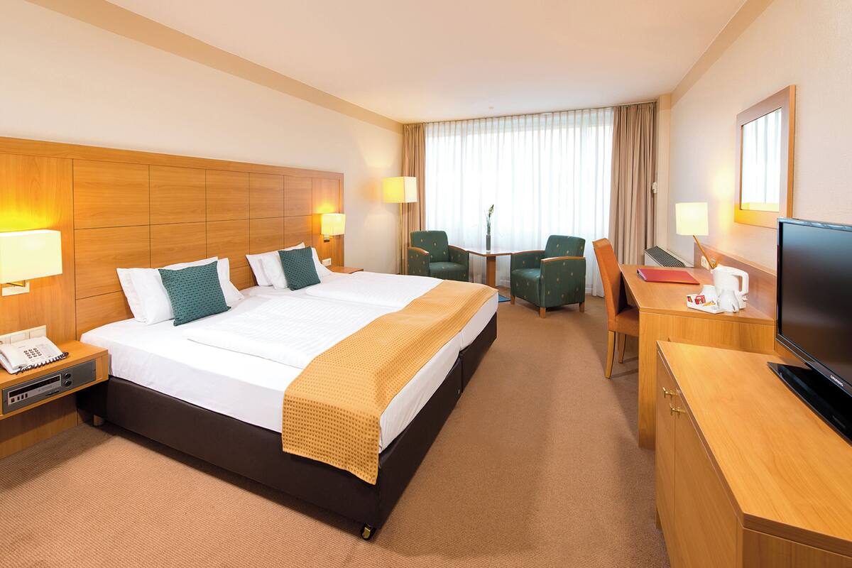 Ein Hotelzimmer mit einem großen Bett, zwei Nachttischen, einem Schreibtisch mit Stuhl, einem Fernseher und einem Fenster mit Vorhängen.