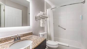Une salle de bain avec un lavabo surmonté d’un plan de travail en granit, des toilettes blanches, une douche en verre et un porte-serviettes.
