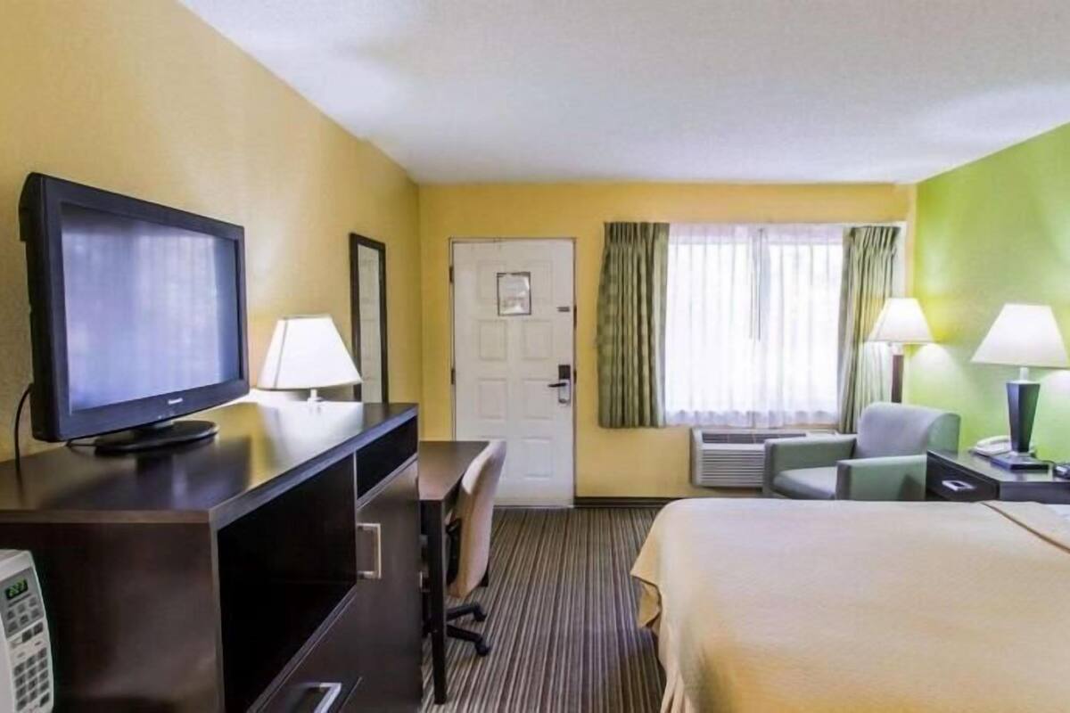 A hotel room with a bed, a desk with a television, a chair, a lamp, and a door.