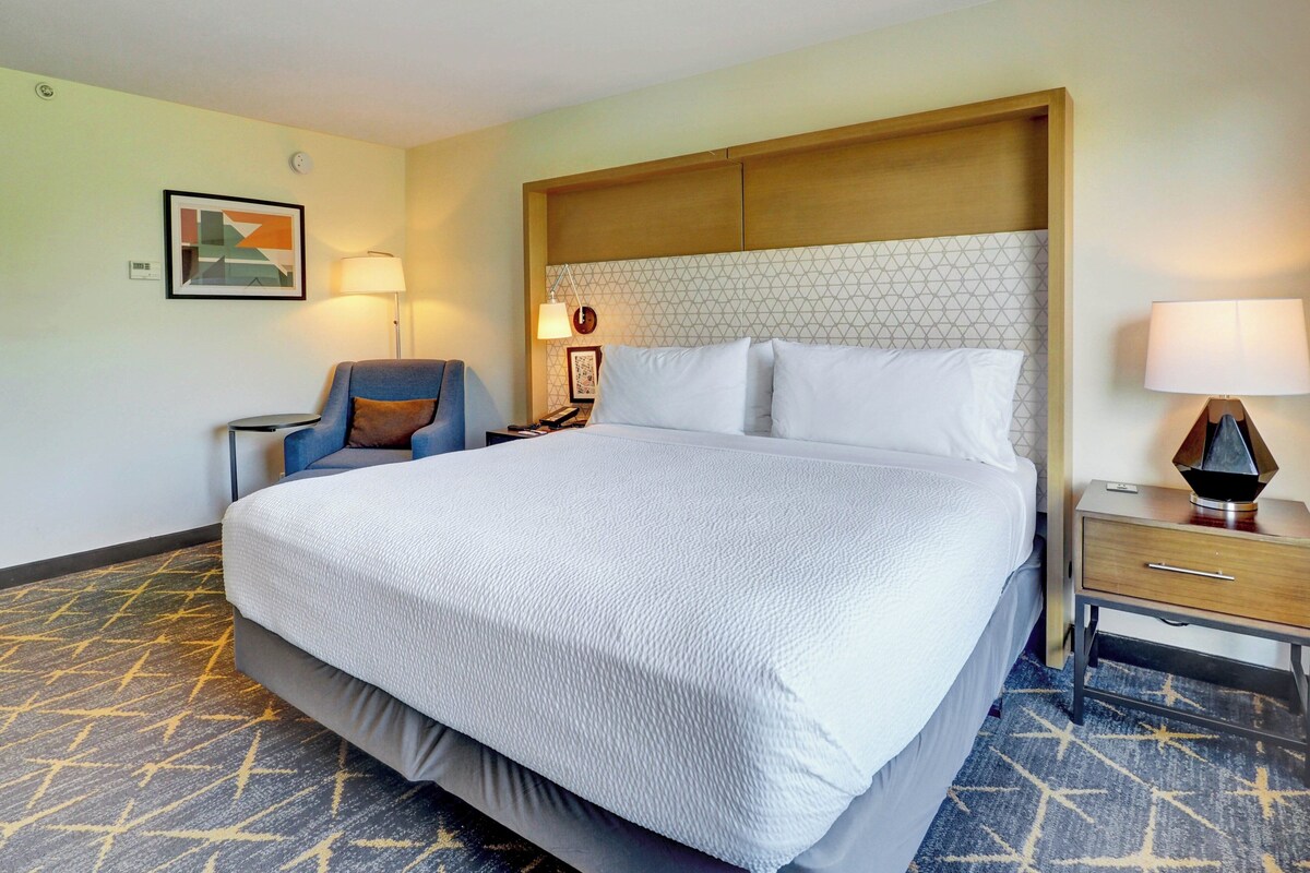 A hotel room with a large bed, a chair, a nightstand, and a wall-mounted lamp.