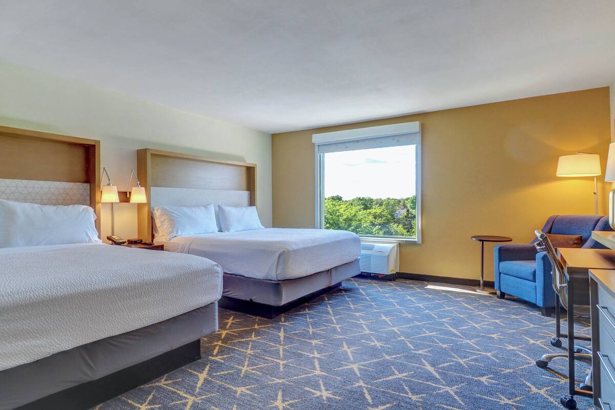 A hotel room with two beds, a desk, a chair, and a window with a view of trees.