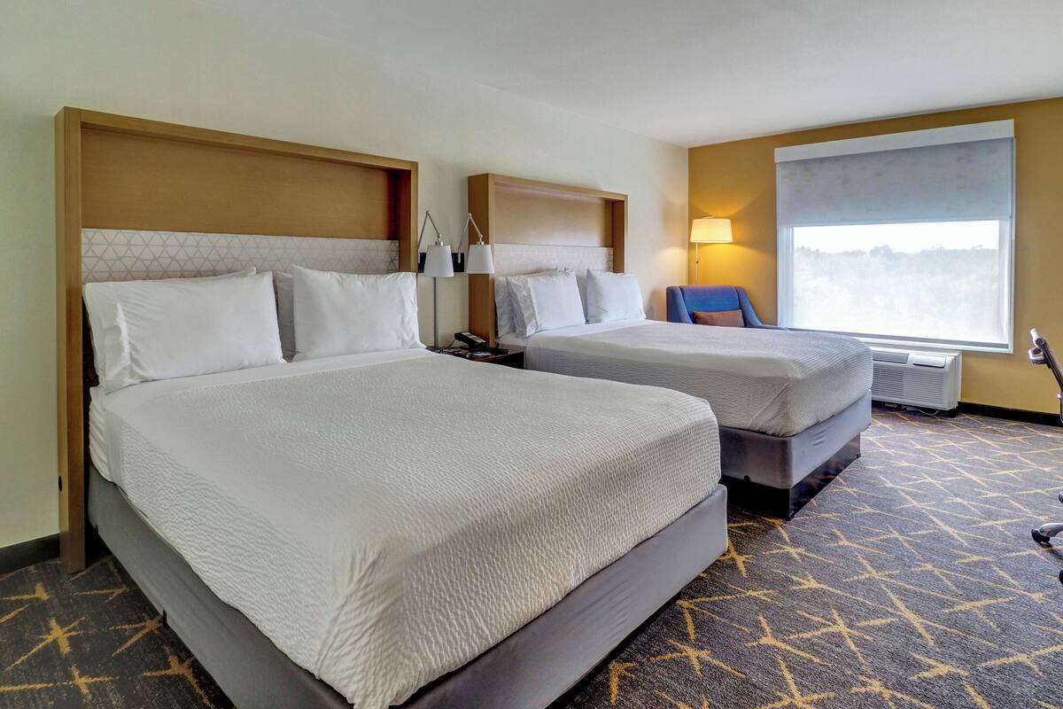 A hotel room with two beds, a nightstand with lamps, a chair, and a large window.