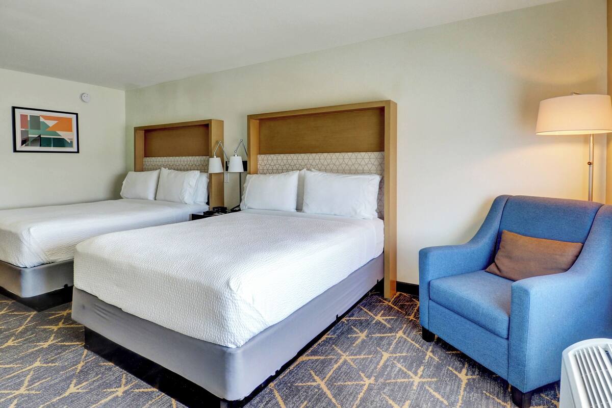 A hotel room with two beds, a blue armchair, a lamp, and a geometric-patterned carpet.