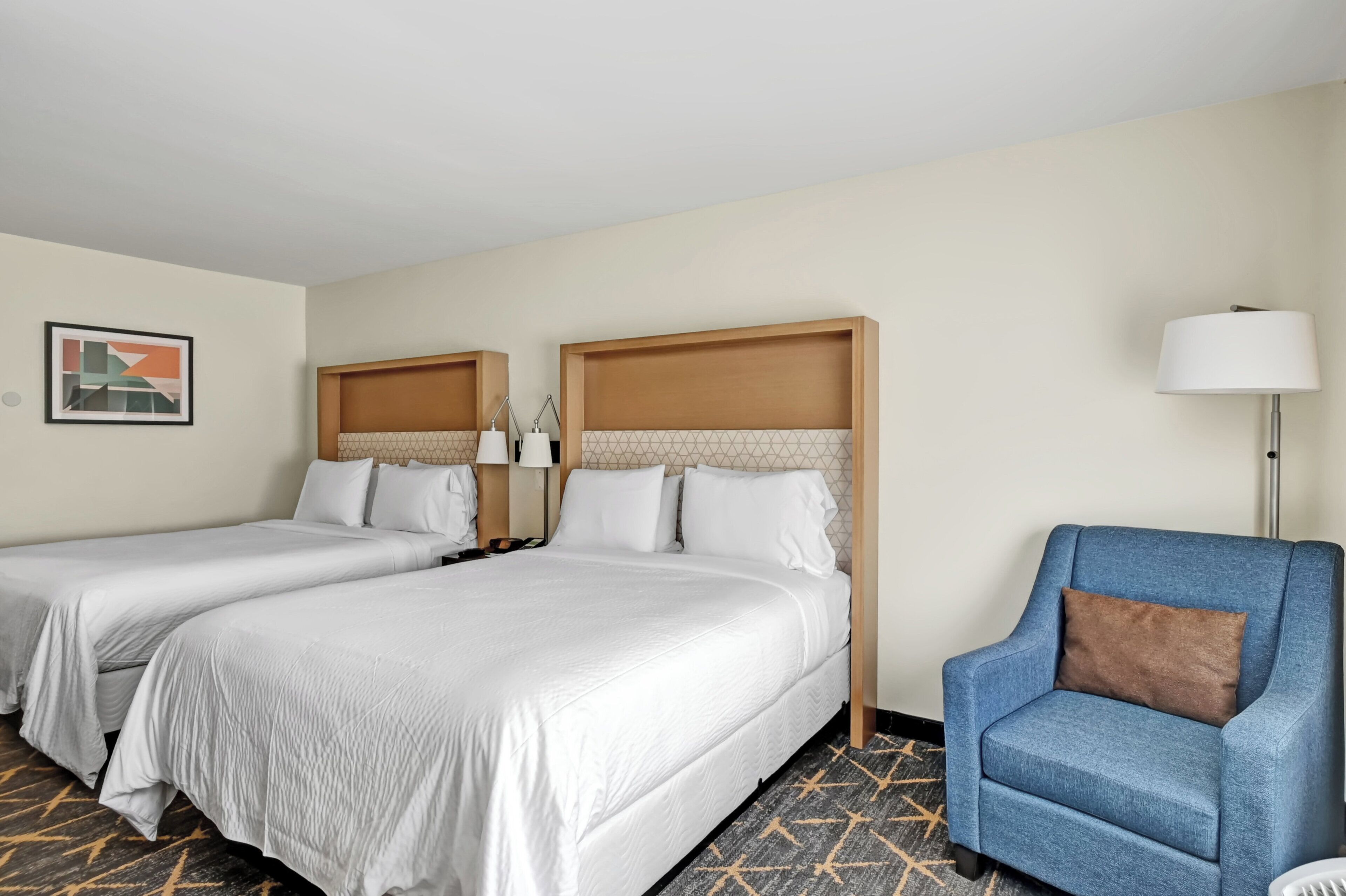A hotel room with two beds, a blue armchair, a lamp, and a framed picture on the wall.