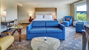 A hotel room with a bed, two blue sofas, a glass coffee table, a desk with a lamp, and a window with a view. - Holiday Inn Piscataway Somerset by IHG (Piscataway)