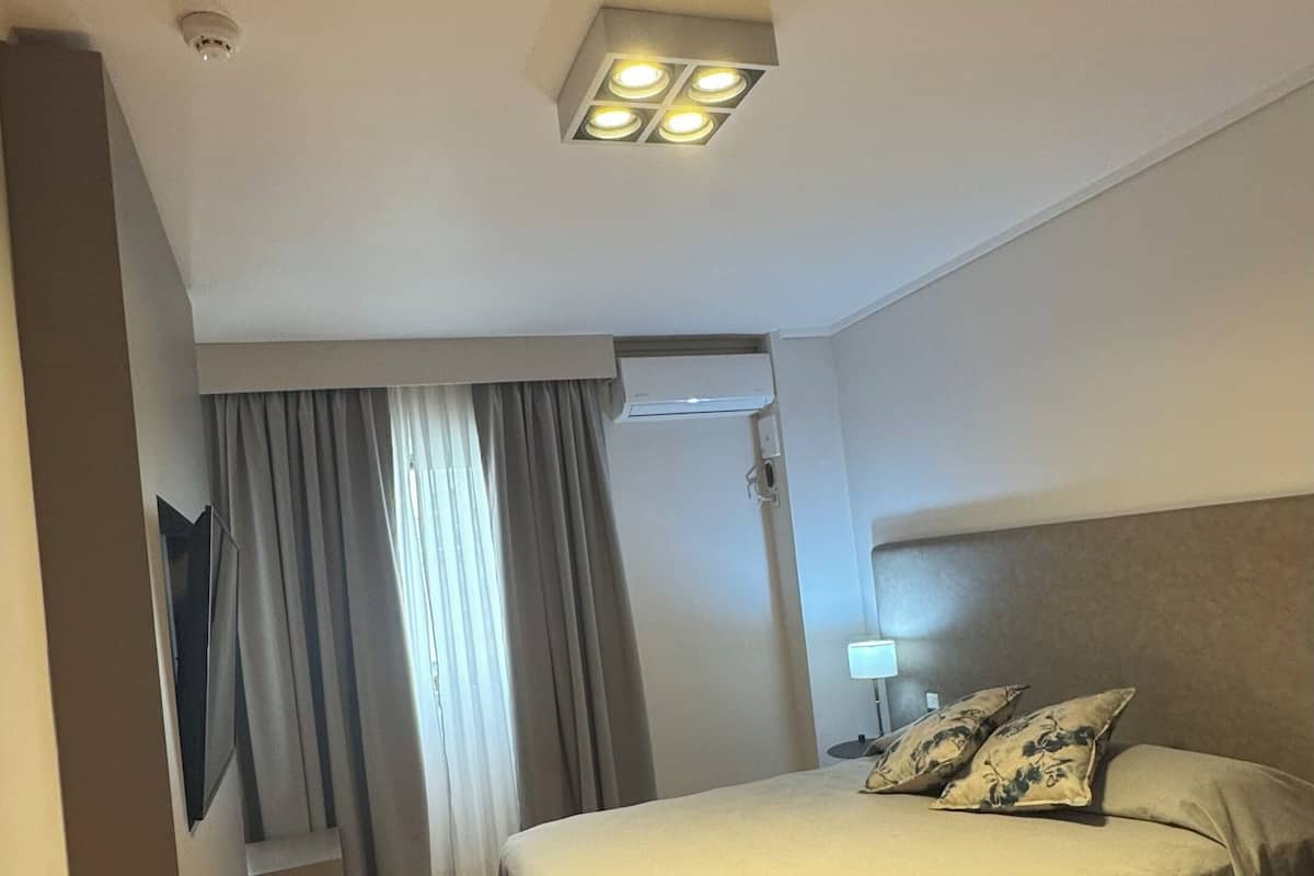Une chambre d’hôtel avec un lit, une table de chevet, une lampe, un luminaire au plafond et une fenêtre avec des rideaux.
