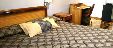 A neatly made bed with patterned bedding, a bedside table, a chair, a desk, and a mirror on the wall.