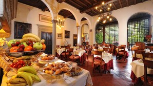 Uma sala de jantar com mesas preparadas para refeições, uma mesa de frutas e um lustre.