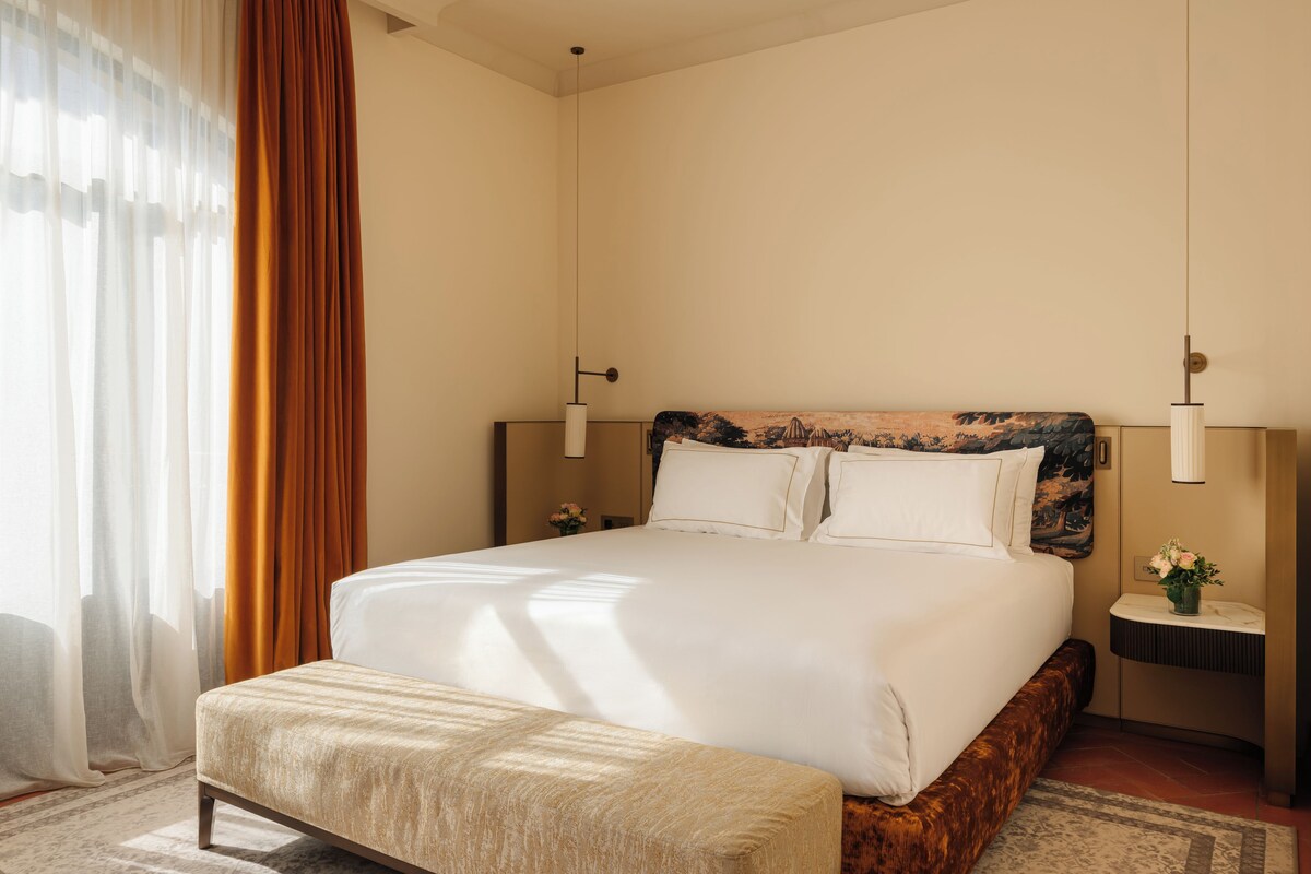 Een hotelkamer met een groot bed, een nachtkastje met een bloevas en een raam met gordijnen.