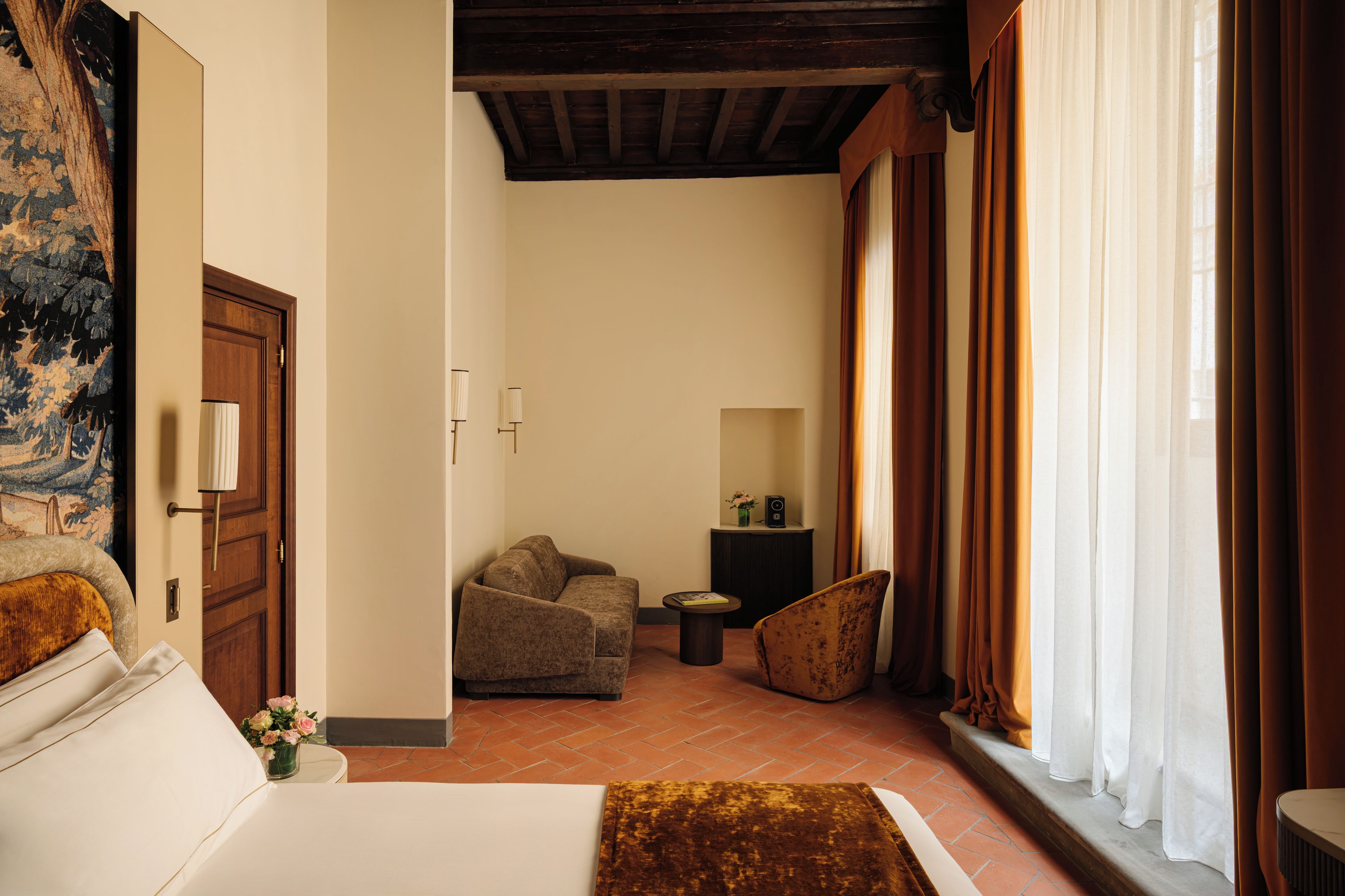 A hotel room with a bed, a sofa, a small table, and a chair. There is a painting on the wall, a door, and a window with curtains.