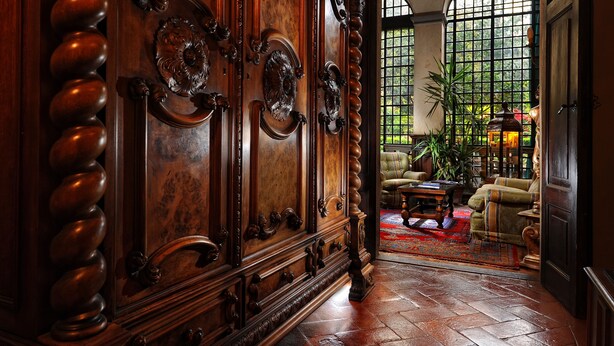 Uma porta de madeira imponente com esculturas detalhadas, uma poltrona confortável, uma mesa de centro e uma janela grande com vista para o verde.