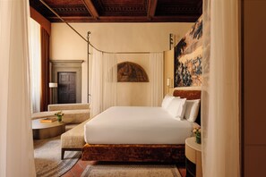 Habitación de hotel con una cama grande, una mesita, una silla, un cuadro en la pared y una puerta.