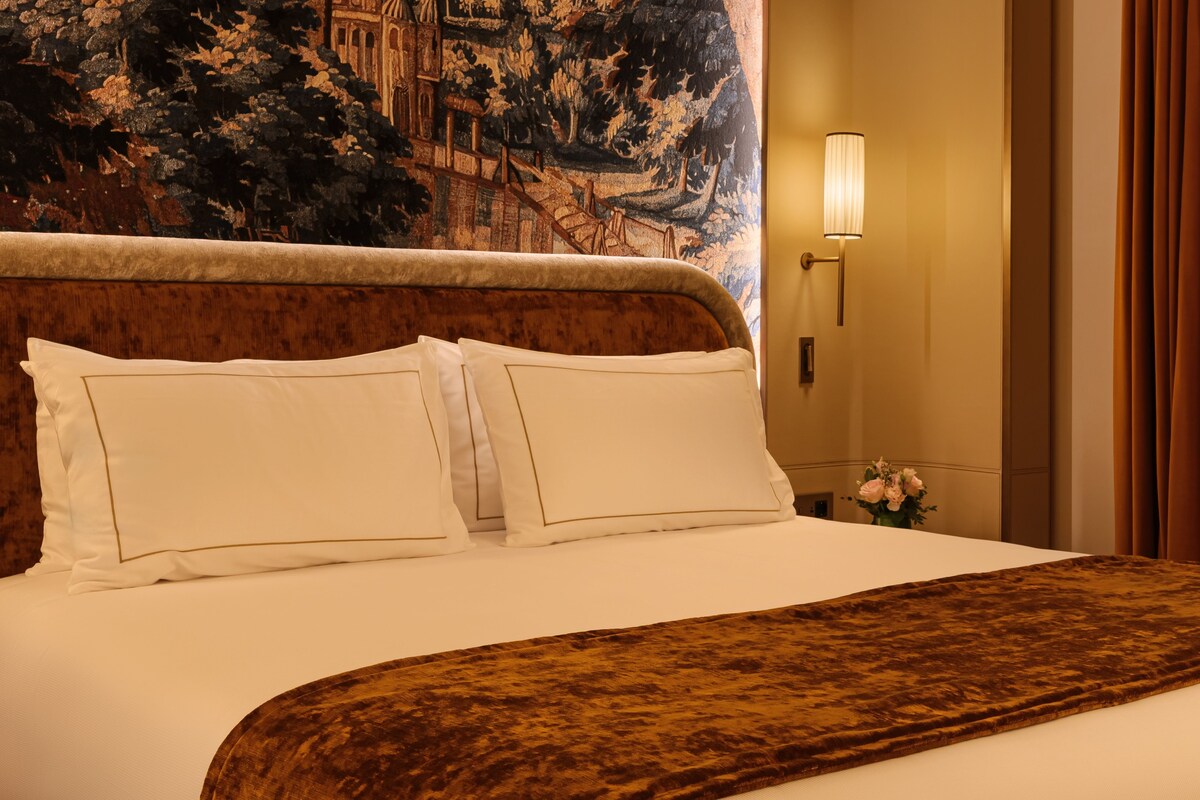 Een hotelkamer met een groot bed, een hoofdbord met een gedetailleerde muurschildering, een nachtkastje met een lamp en een bloemstuk.