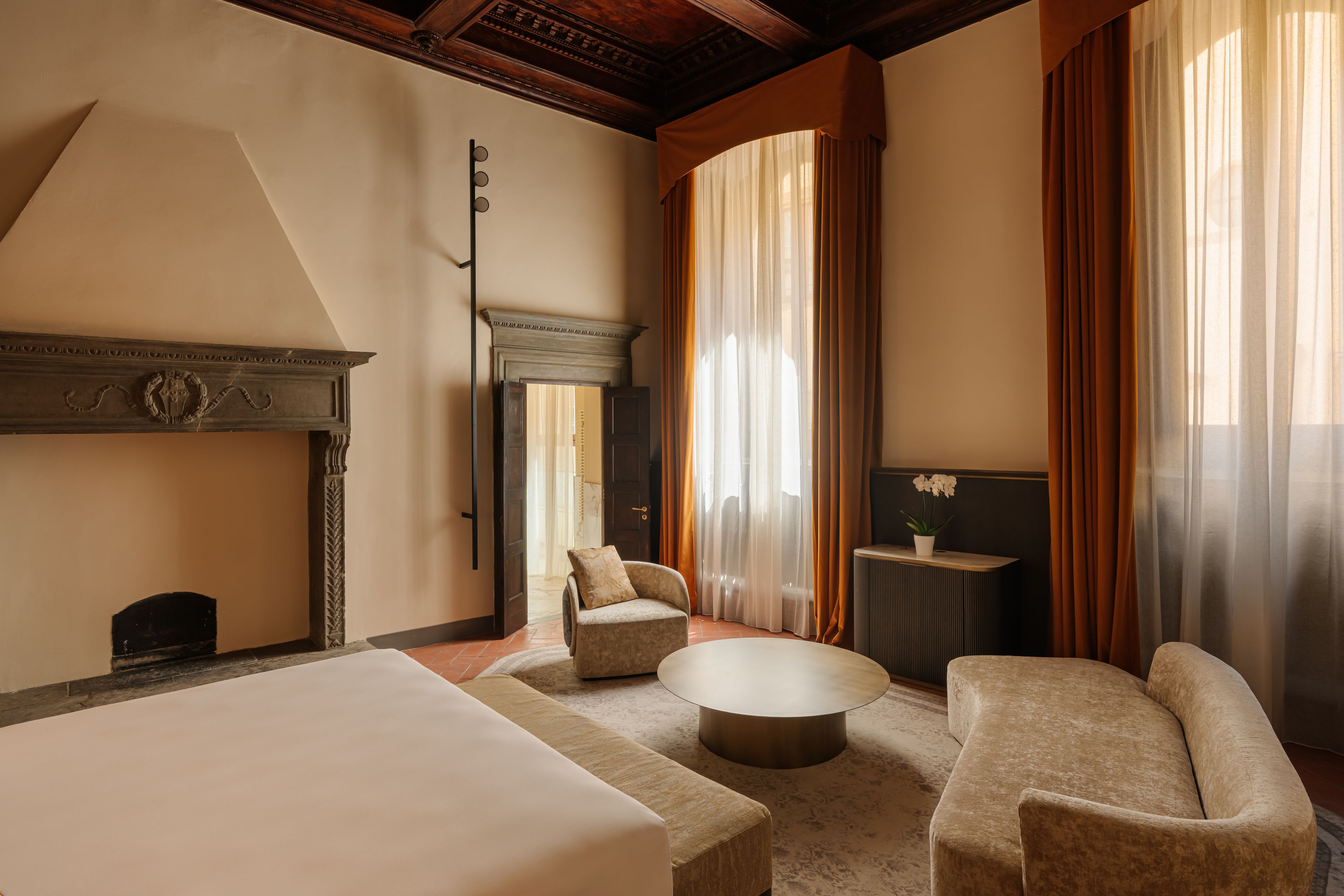 A hotel room with a large bed, two armchairs, a round coffee table, a fireplace, and a view of the outdoors through sheer curtains.