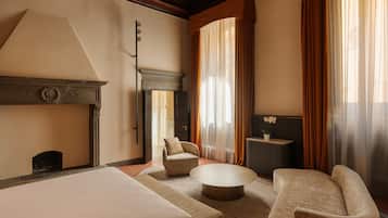 Habitación de hotel con una cama grande, dos sillones, una mesa de centro redonda, una chimenea y vistas al exterior a través de cortinas translúcidas.