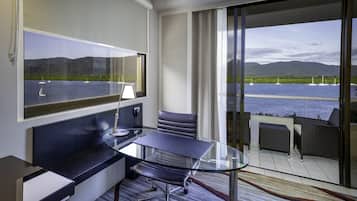Una habitación de hotel moderna con un escritorio de vidrio, sillones de cuero, un ventanal con vistas al mar y a las montañas, y un balcón con mobiliario de mimbre.