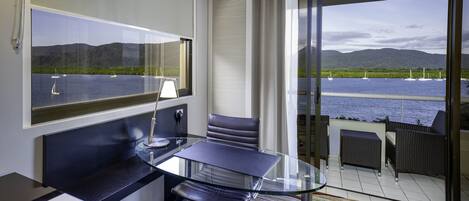 Una habitación de hotel moderna con un escritorio de vidrio, sillones de cuero, un ventanal con vistas al mar y a las montañas, y un balcón con mobiliario de mimbre.