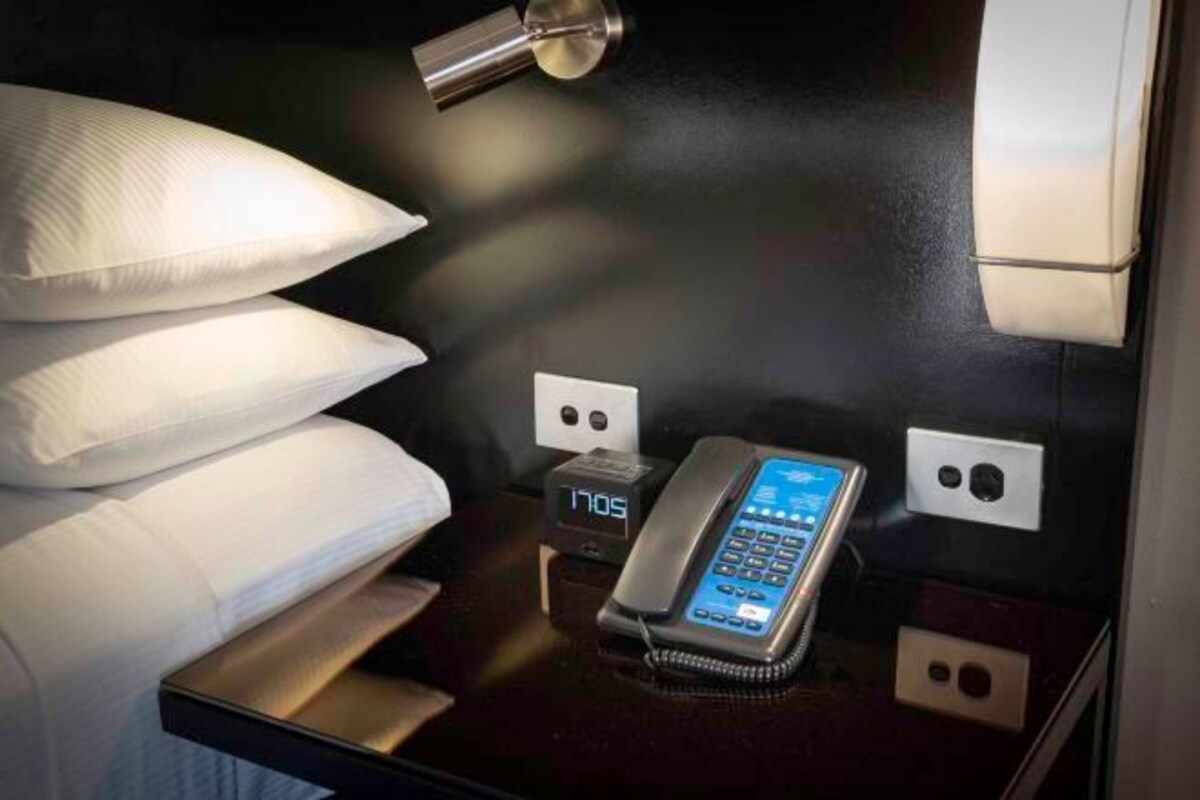 Habitación de hotel con una cama, almohadas, una mesita de noche, un reloj digital y un teléfono.