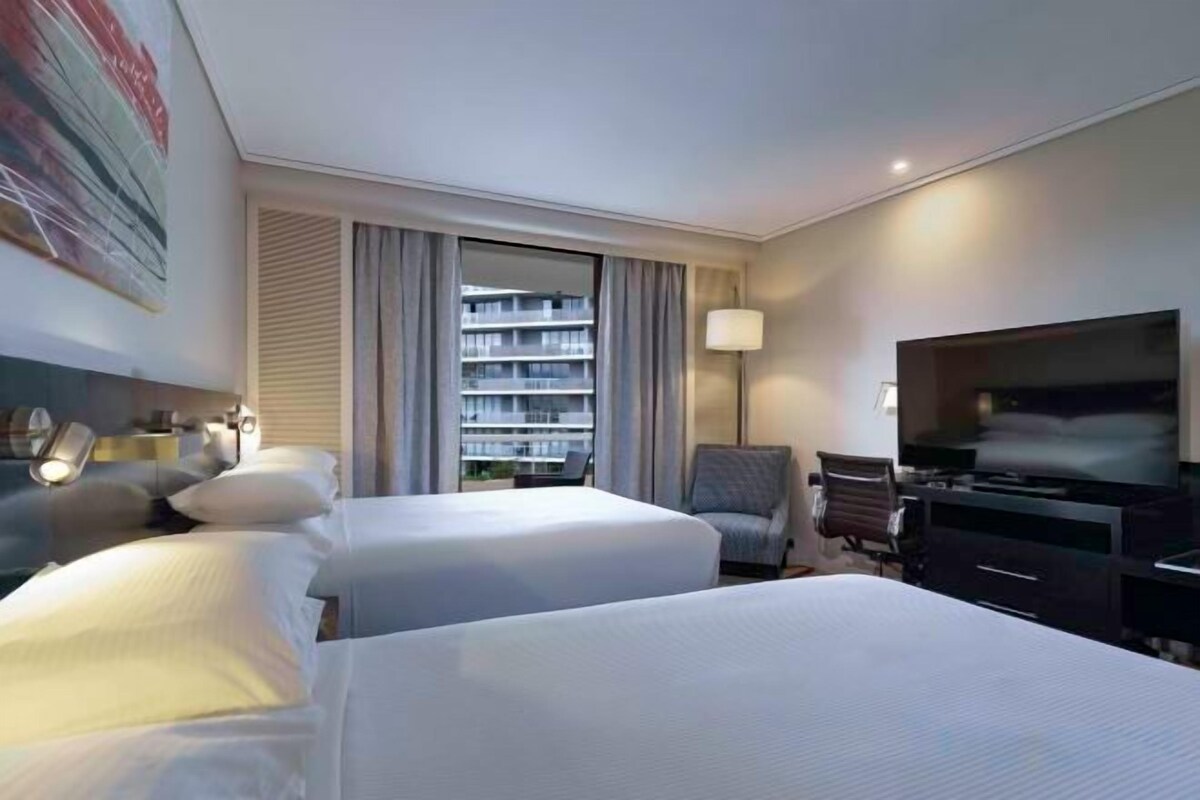 A hotel room with two beds, a TV, a desk, and a window with curtains.