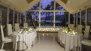 An indoor dining area with tables set for a meal, large windows with a view of palm trees and the ocean, and a decorative harp in the center. - Hilton Cairns (Cairns)