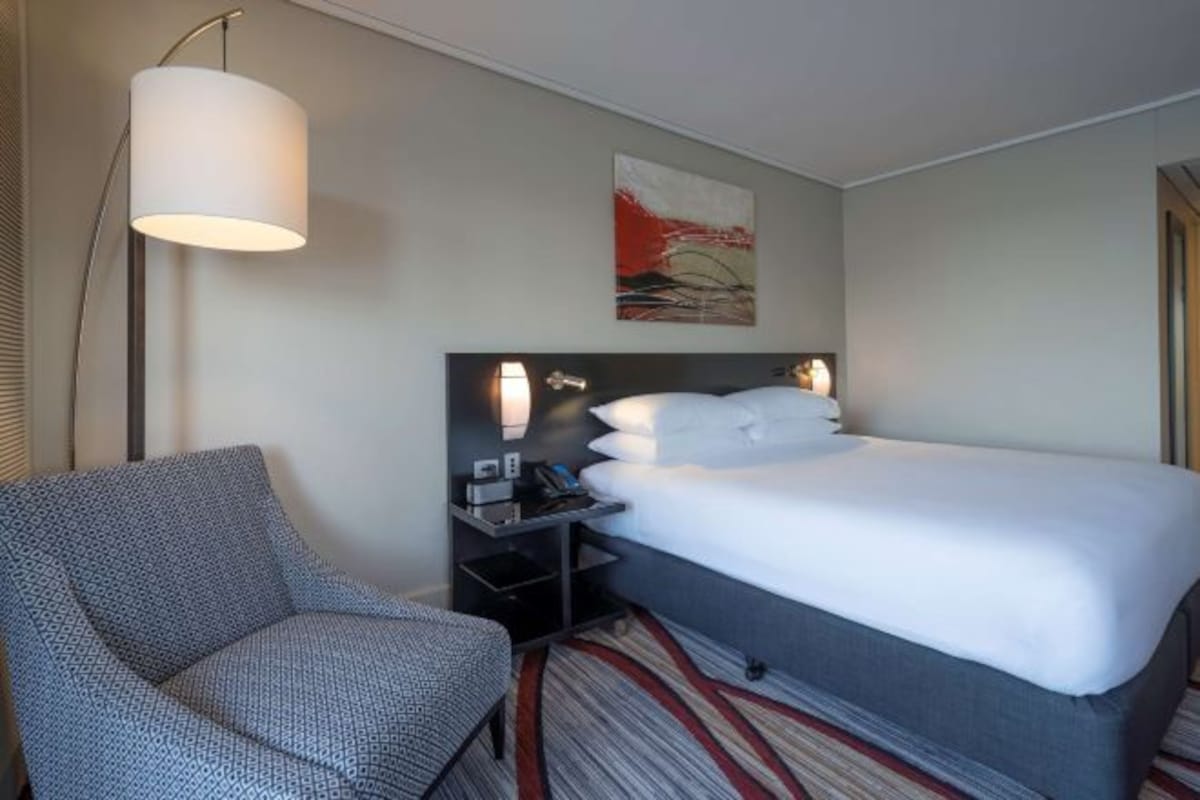 A hotel room with a bed, a chair, a bedside table with a lamp, and a painting on the wall.