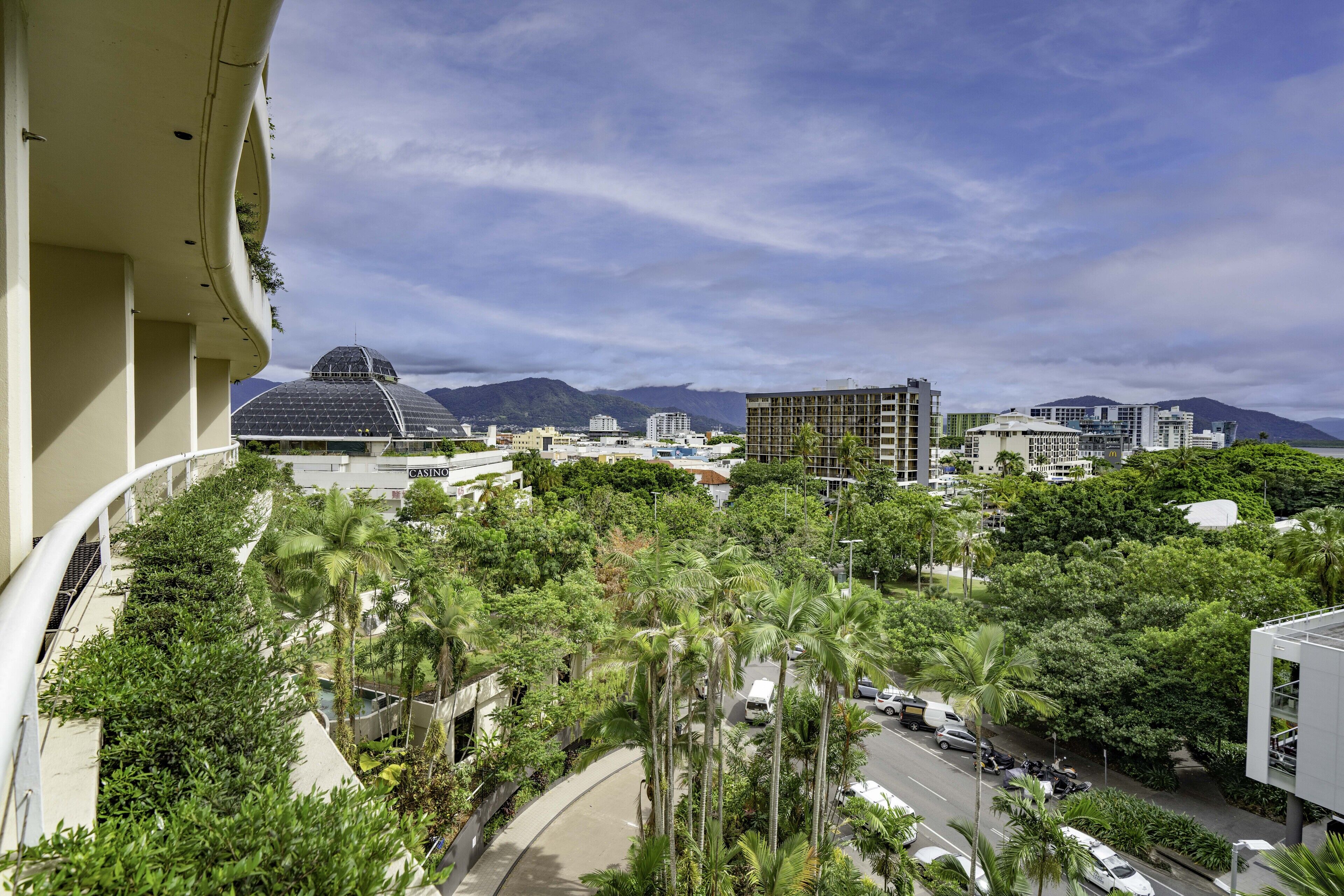 A view from a balcony overlooking a cityscape with buildings, palm trees, and mountains in the background.