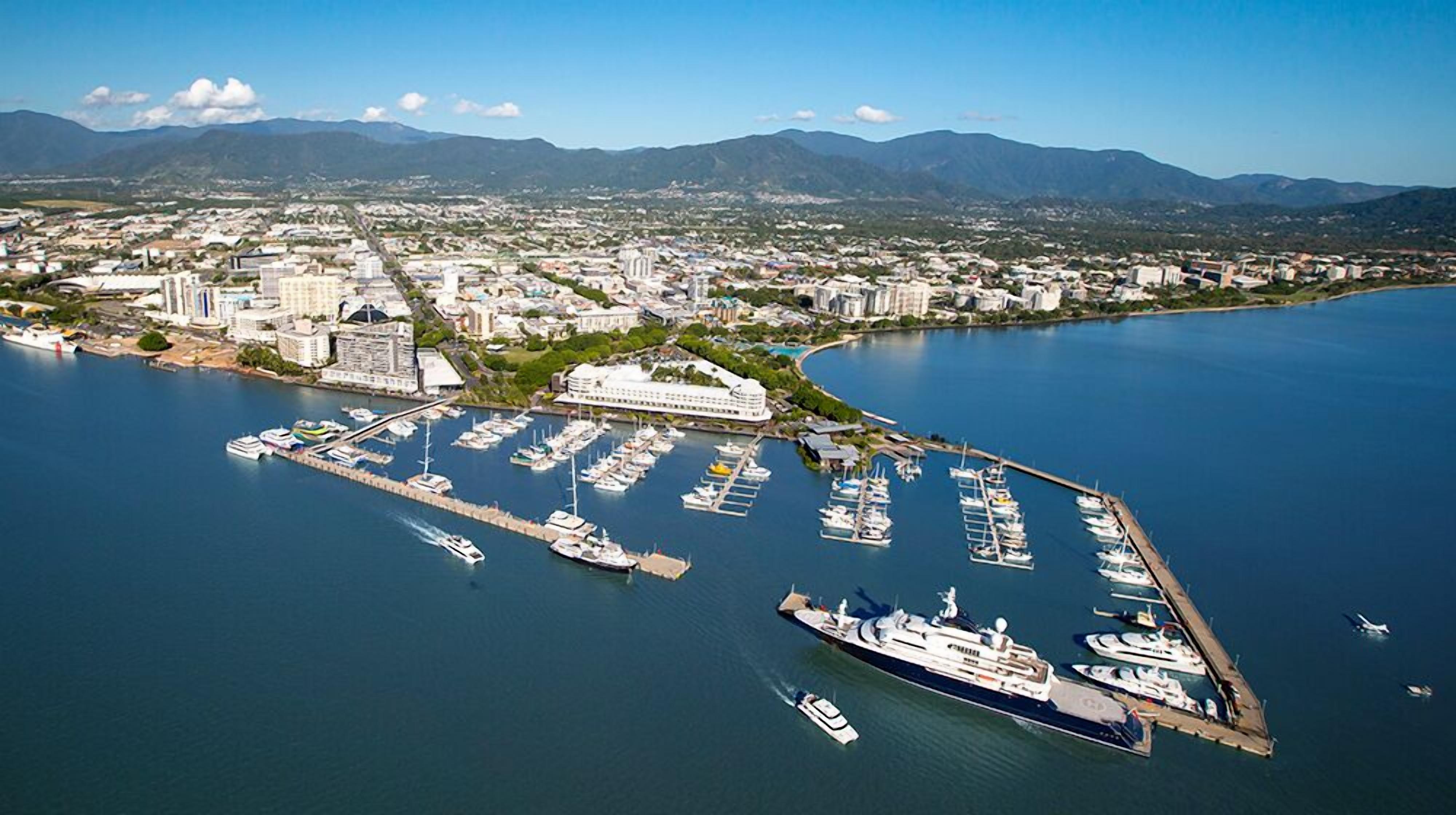 An aerial view of a marina with boats docked, a large cruise ship, and a cityscape with mountains in the background.