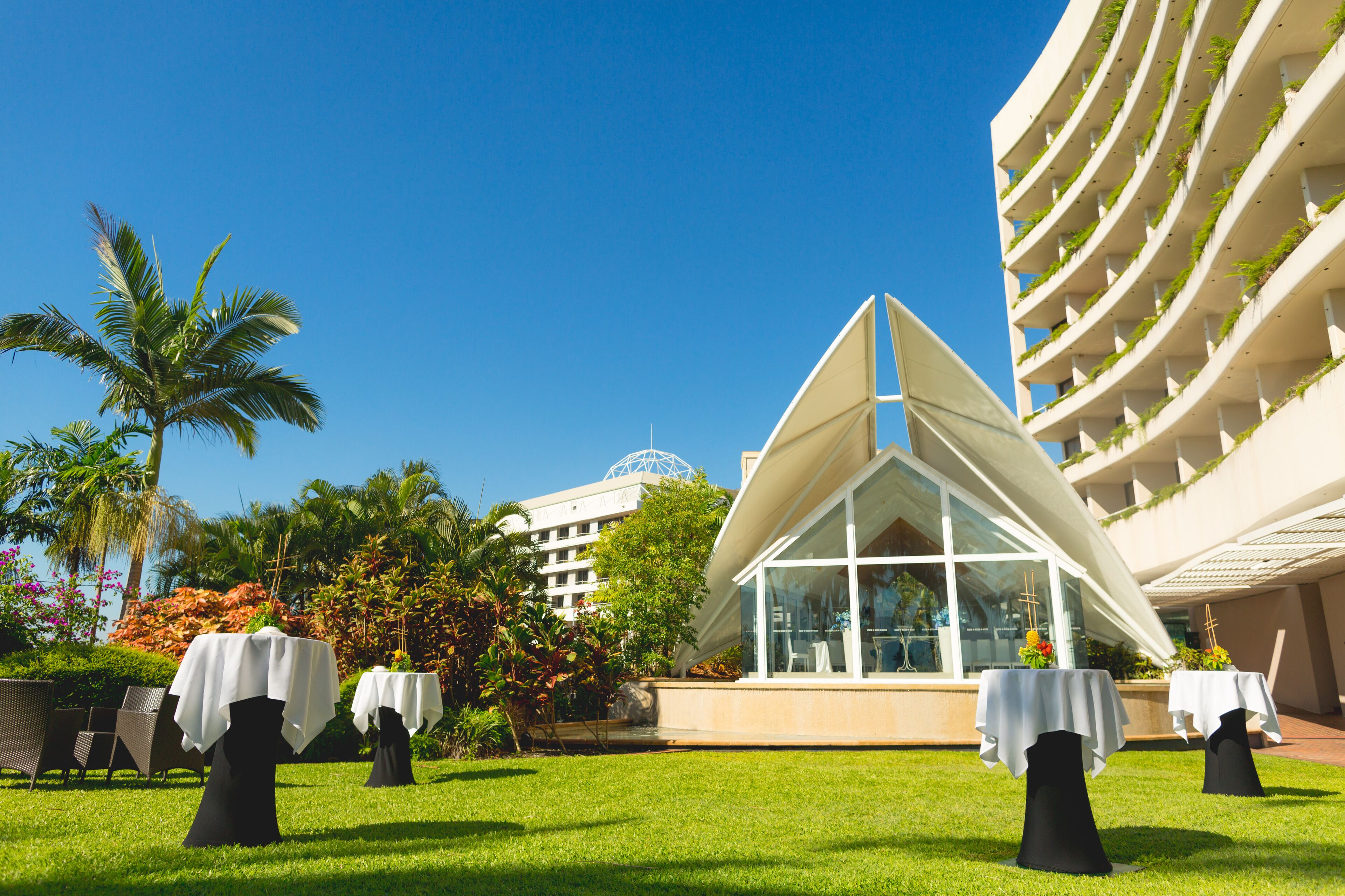 A hotel with outdoor dining tables set up on a lawn, surrounded by lush greenery and palm trees.