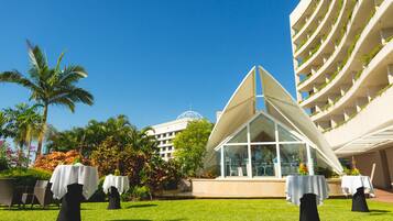A hotel with outdoor dining tables set up on a lawn, surrounded by lush greenery and palm trees.