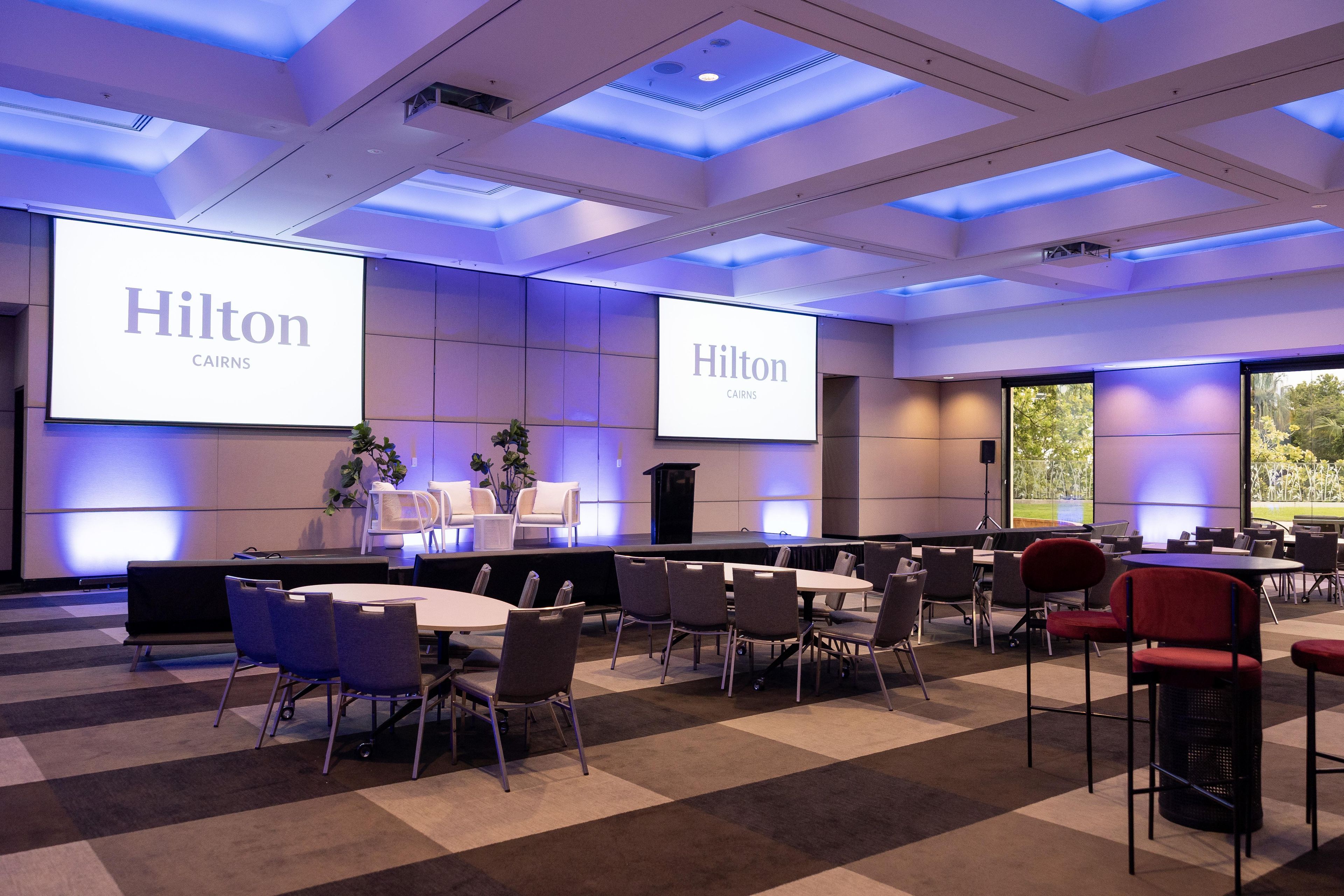 Ein Konferenzraum mit einer Bühne, mehreren Tischen und Stühlen, ausgestattet mit großen Bildschirmen, die 'Hilton' und 'CAIRNS' zeigen.