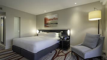 A modern hotel room with a large bed, a chair, a nightstand with a lamp, and a painting on the wall.