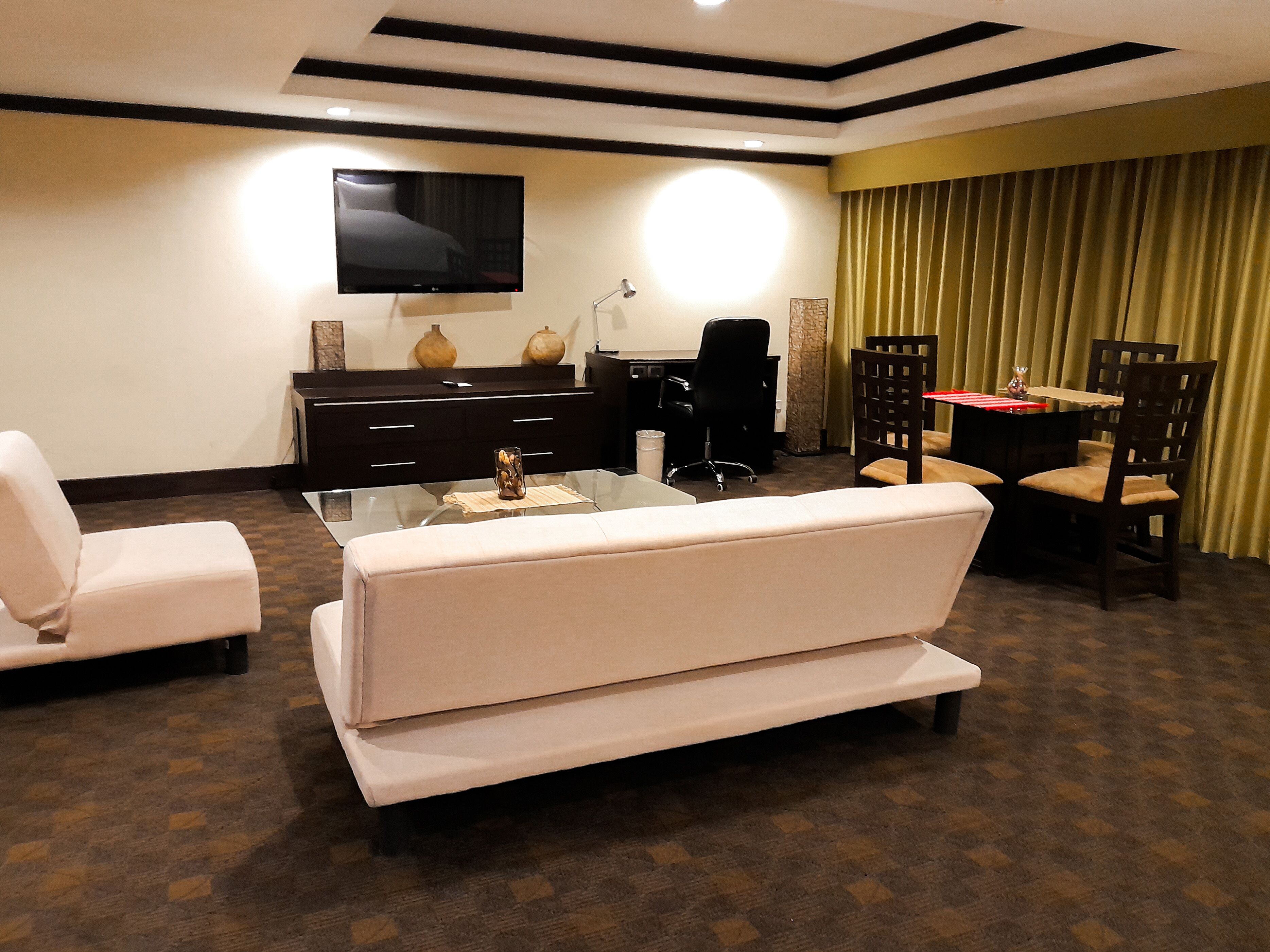 A modern living room with a sofa, a glass coffee table, a TV mounted on the wall, a dining table with chairs, and a patterned carpet.