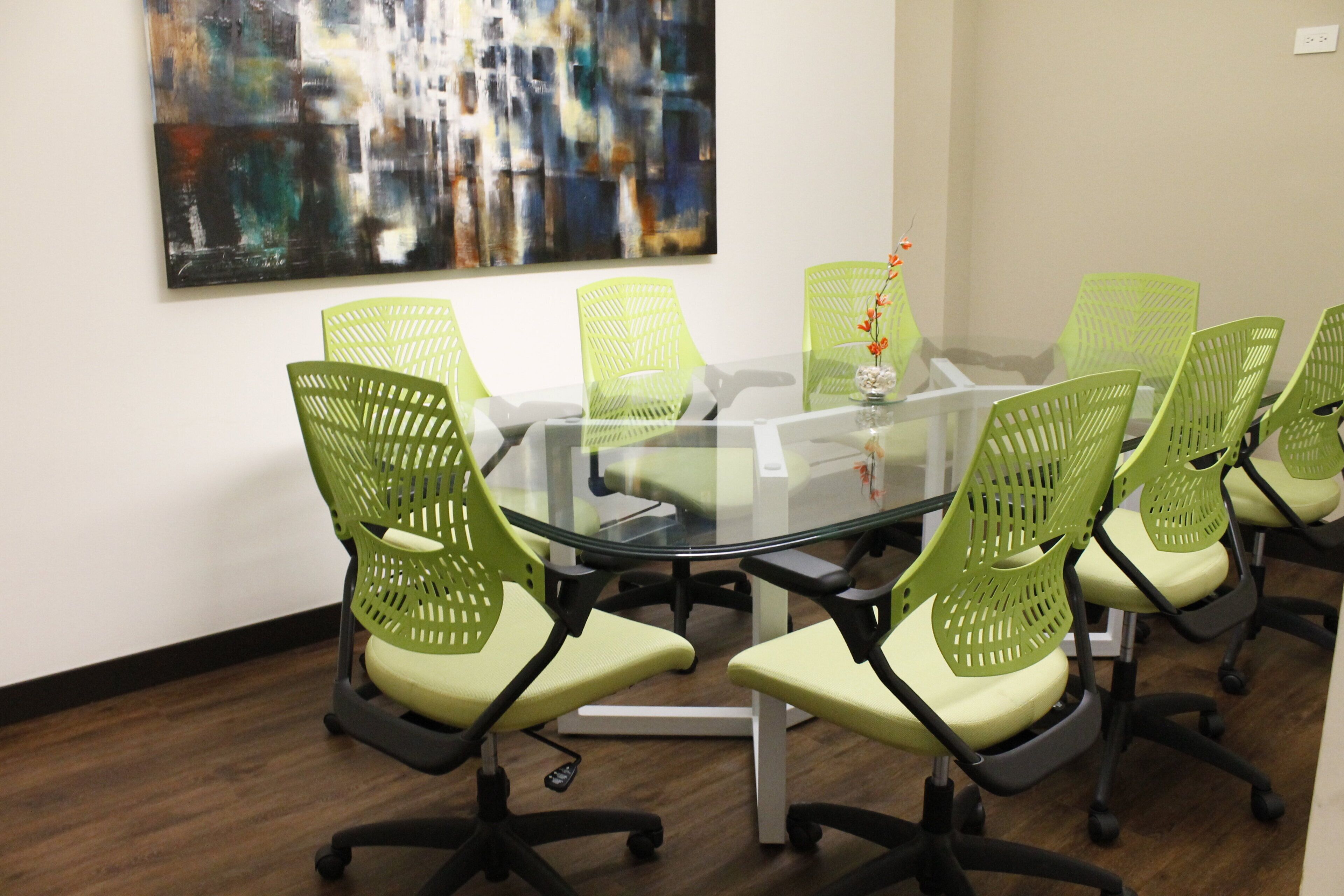 A conference room with a glass table surrounded by green chairs with black frames and black wheels.