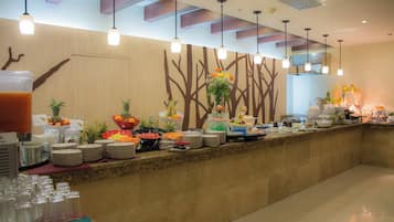 A buffet setup with various food items and beverages, a wall with a tree design, and hanging pendant lights.