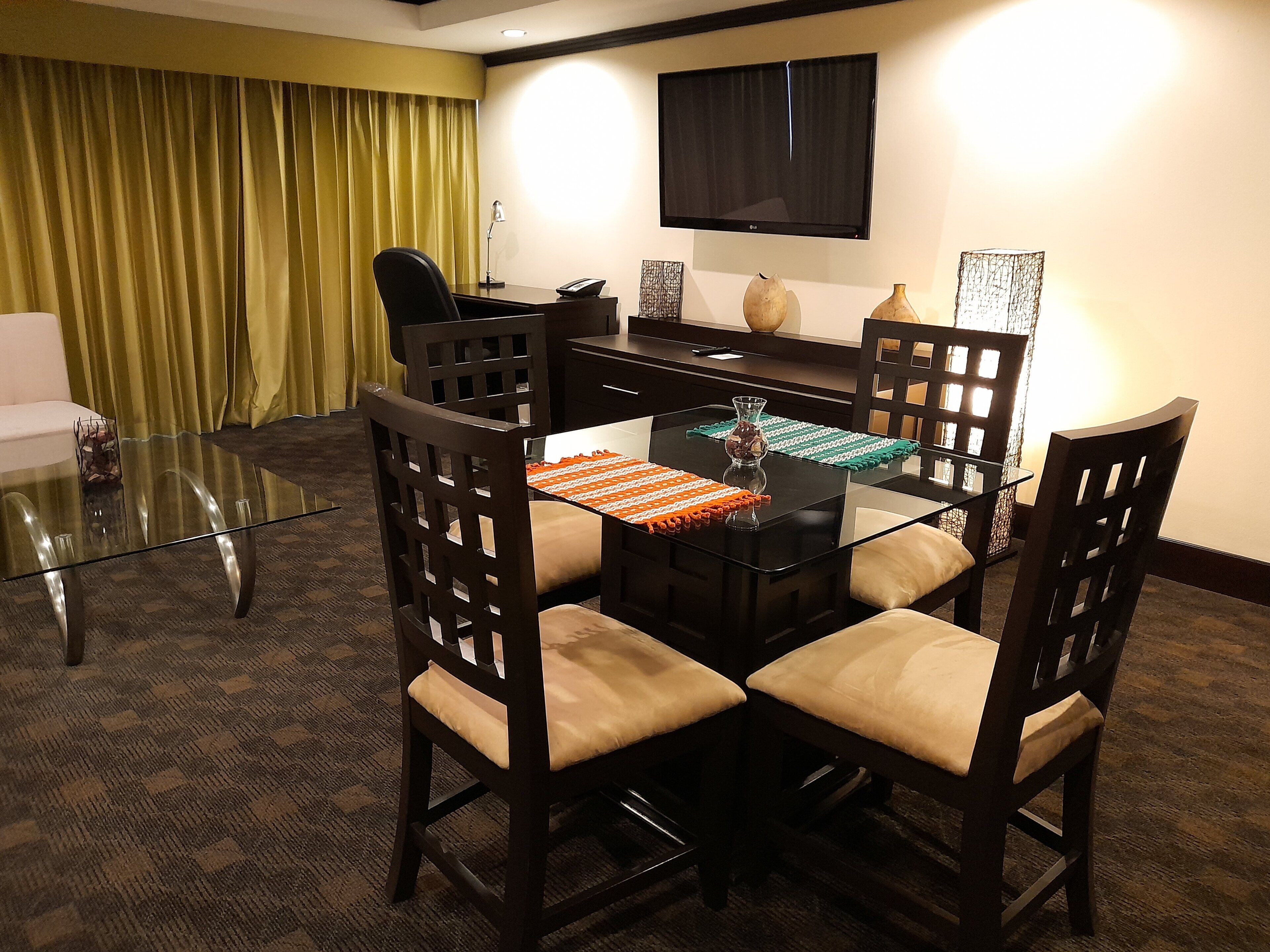 A conference room with a glass table, four wooden chairs, a TV mounted on the wall, and a yellow curtain.