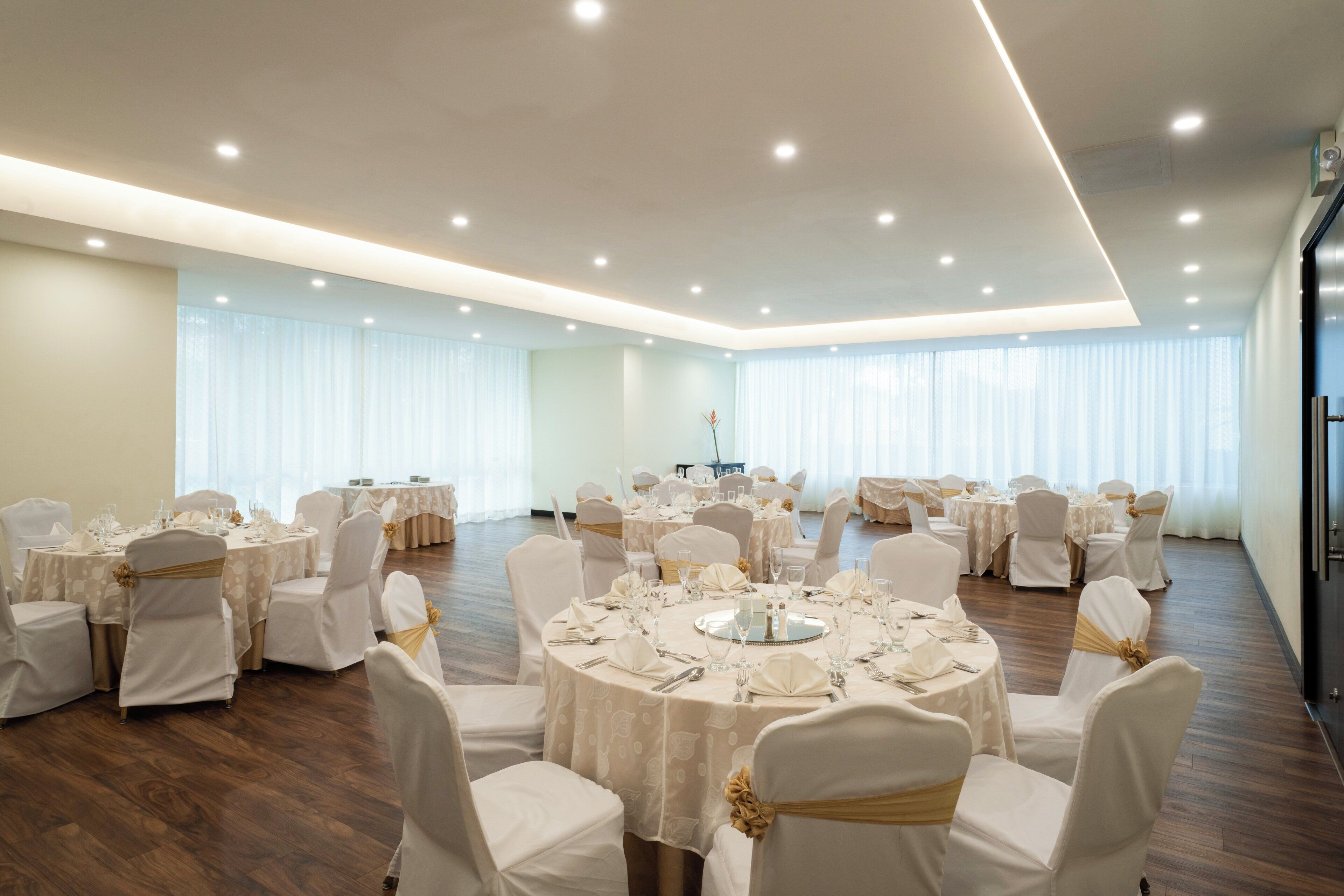 Un amplio salón de banquetes con mesas redondas, sillas blancas y cintas doradas, que presenta un diseño moderno en el techo y ventanas grandes con cortinas translúcidas.
