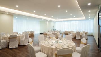 Un amplio salón de banquetes con mesas redondas, sillas blancas y cintas doradas, que presenta un diseño moderno en el techo y ventanas grandes con cortinas translúcidas.