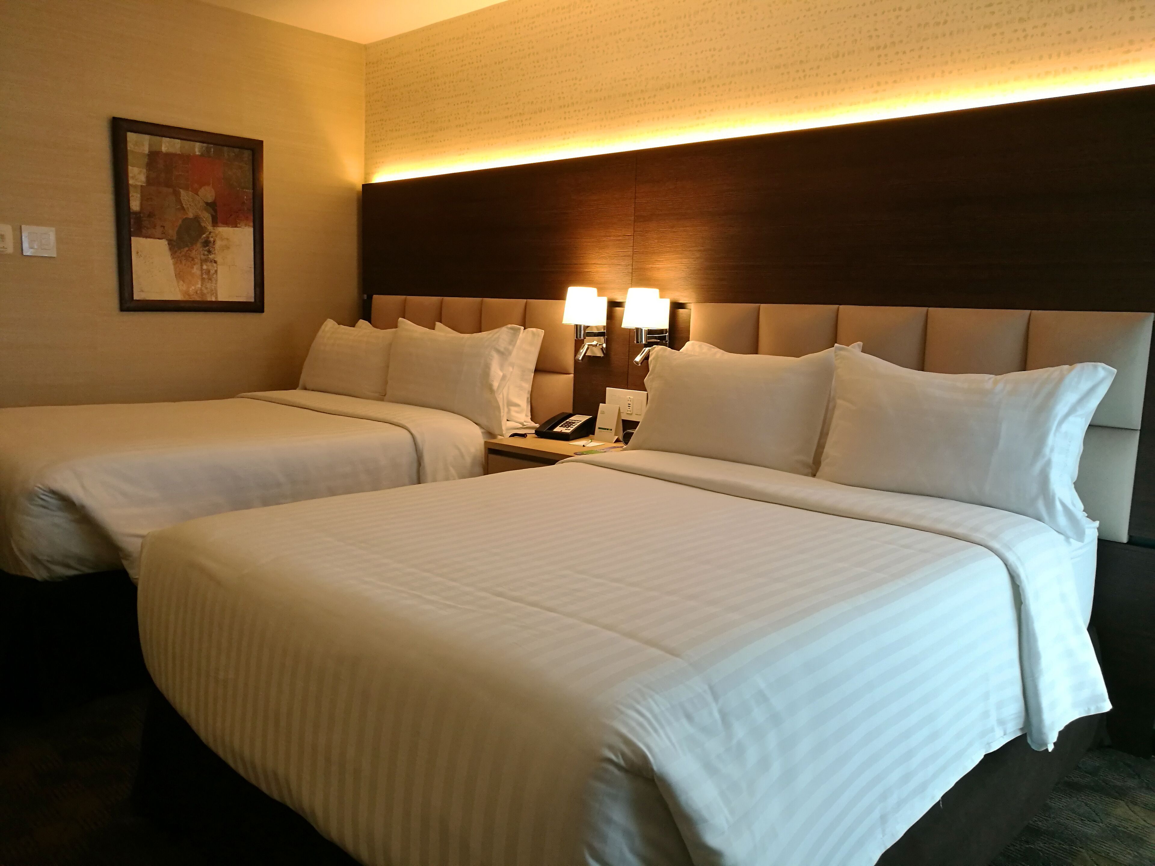 A hotel room with two beds, a headboard with a light fixture, a nightstand with a phone and a lamp, and a framed picture on the wall.