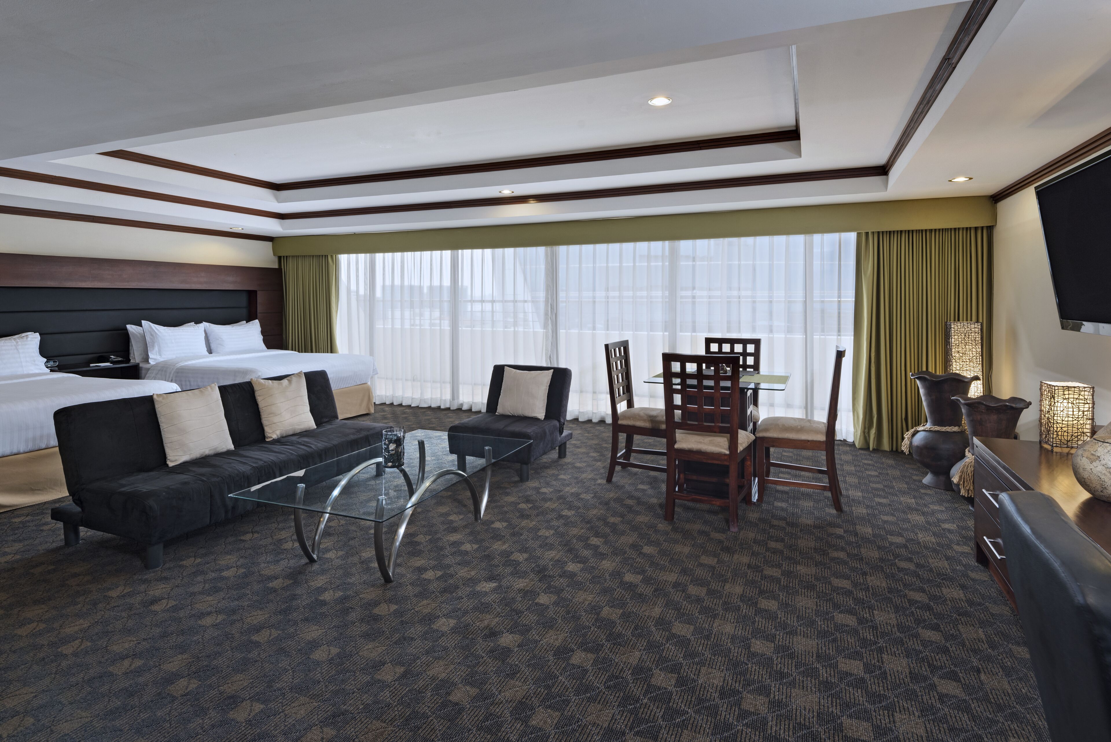 Habitación de hotel con cama, sofá, mesa de centro de vidrio, mesa de comedor con sillas y televisor en la pared.