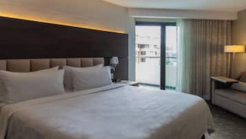 A hotel room with a large bed, a sofa, a nightstand, and a view of the city through a large window.
