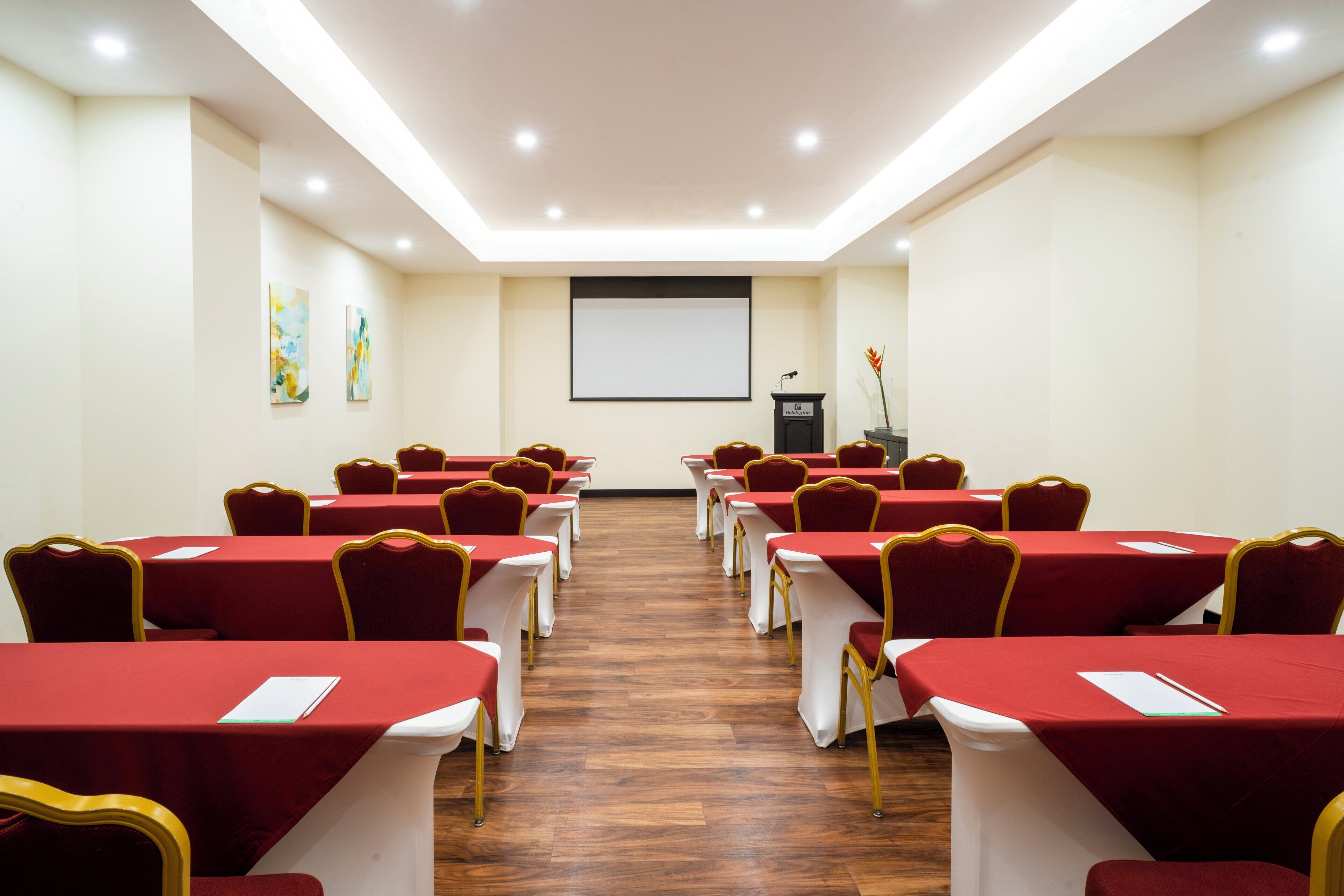 A conference room with rows of tables and chairs, a podium, and a projector screen.