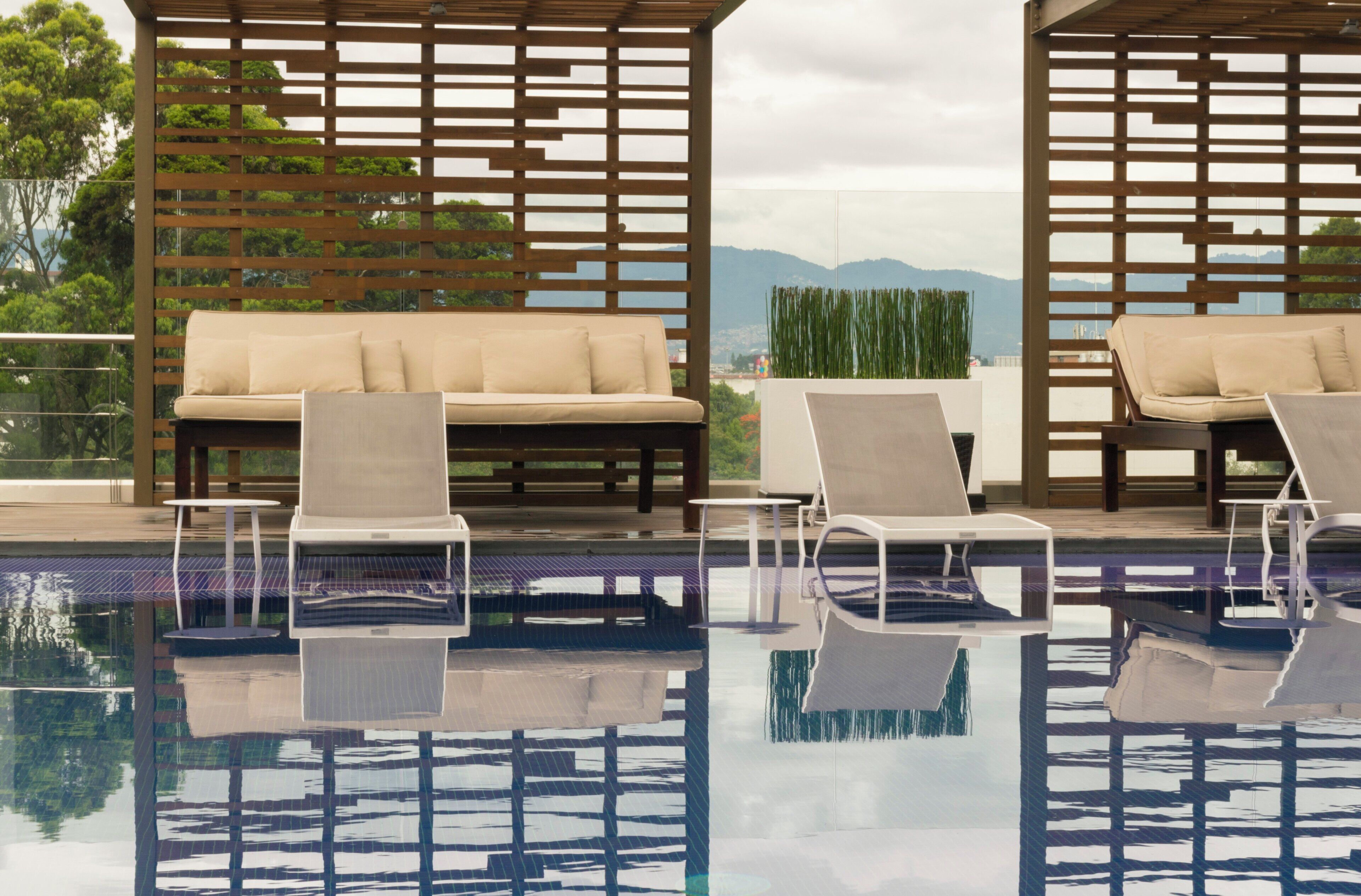 Zona de piscina con sillones de descanso y un sofá, rodeada por estructuras de celosía de madera.