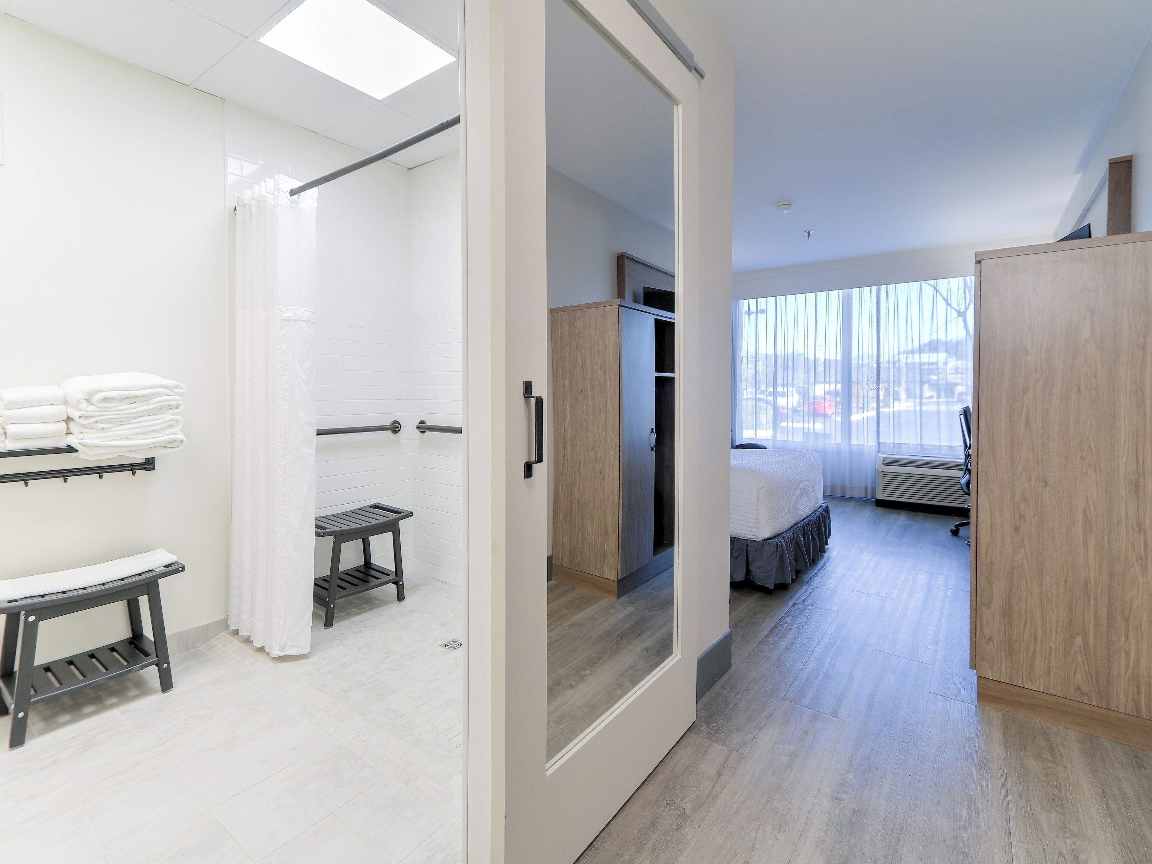 A modern hotel room with a shower, a bed, a wardrobe, and a view of the city.
