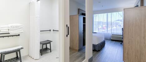 A modern hotel room with a shower, a bed, a wardrobe, and a view of the city.