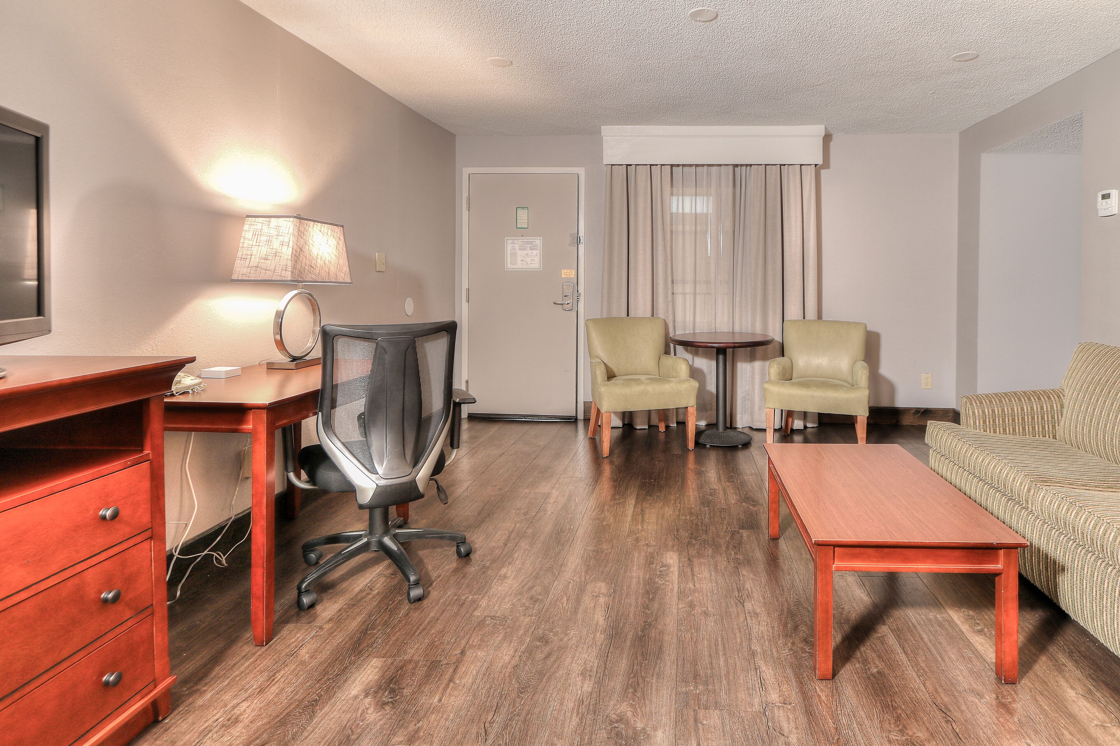 A hotel room with a wooden desk, two armchairs, a sofa, a small table, and a lamp.
