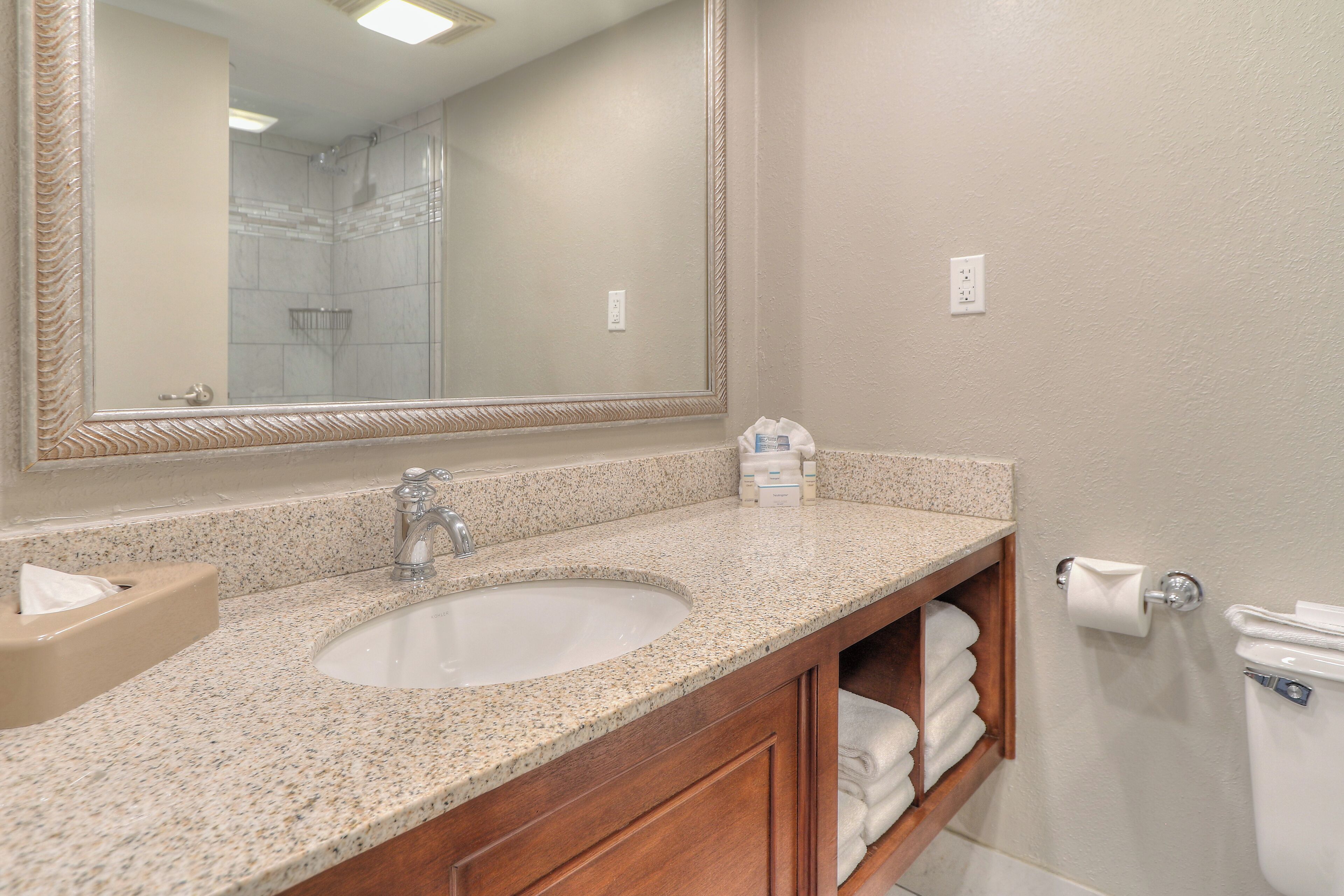 A bathroom with a granite countertop sink, a large mirror, a wooden vanity, and a toilet paper holder.