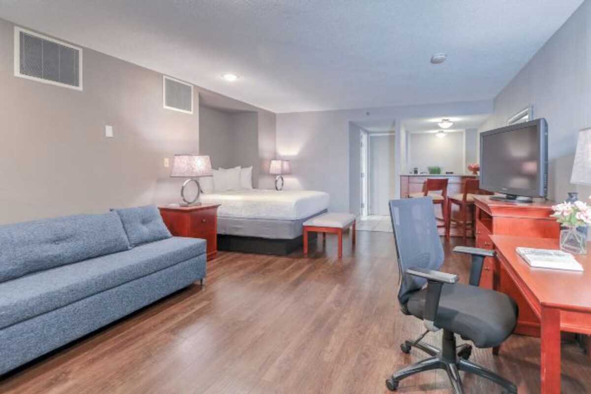 Una habitación de hotel moderna con cama, sofá, escritorio con silla, televisor y una mesita de noche con lámpara.