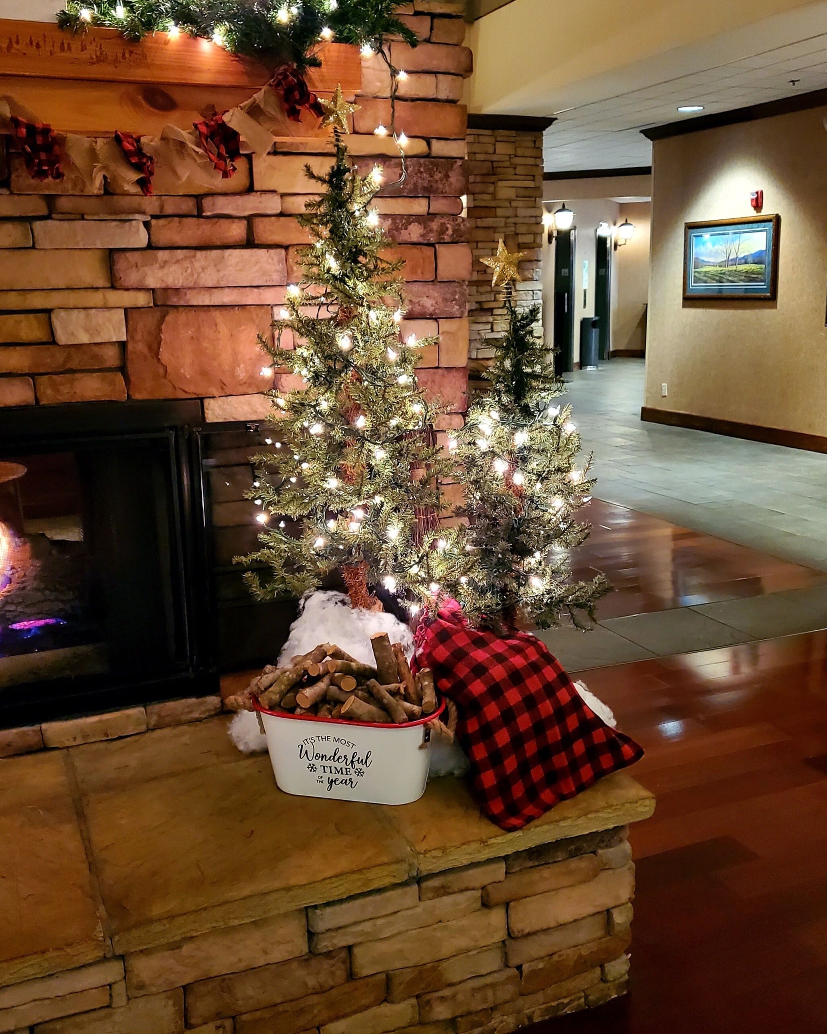 Un árbol de Navidad decorado en un vestíbulo con chimenea, una cesta de leños y un cojín a cuadros.